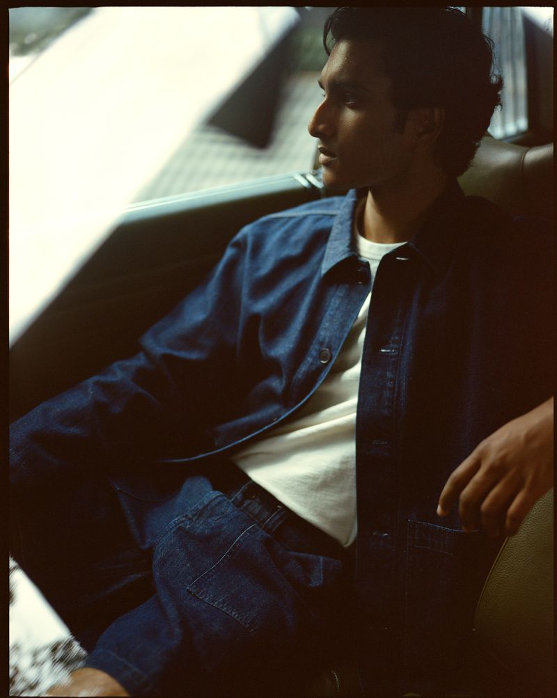 Portrait of a handsome man in denim jacket and pants sitting in a car, looking out the window in soft, diffused light.