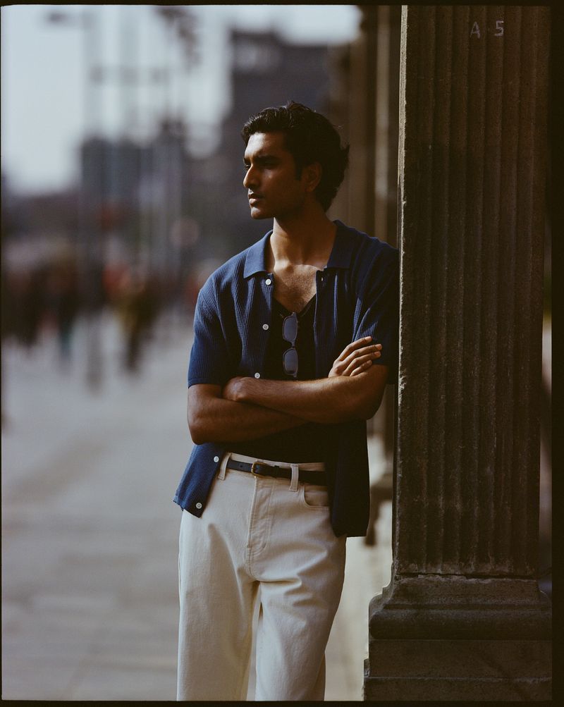 Portrait of a handsome man with dark hair, wearing a navy cardigan and cream pants, leaning against a pillar in an urban setting.