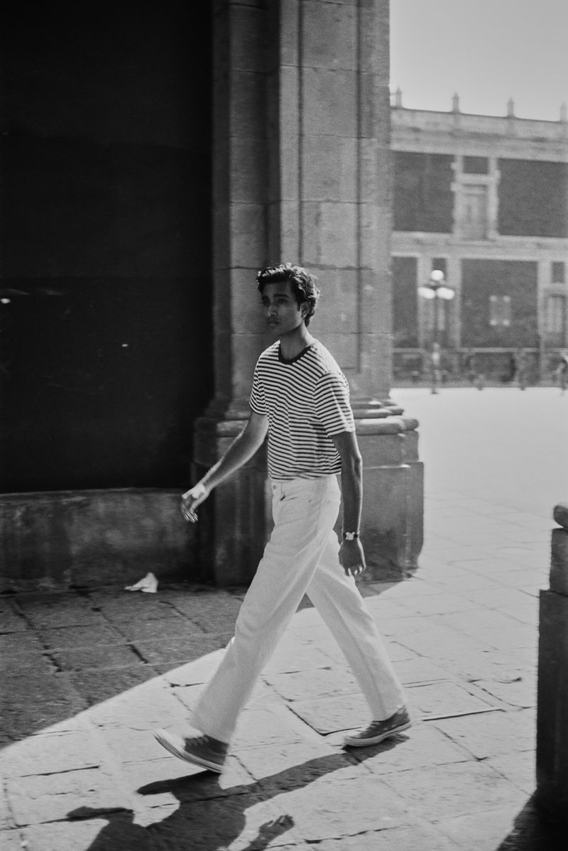 A man walks through an arched stone doorway in monochrome, wearing a striped shirt and white pants in an urban setting with buildings.