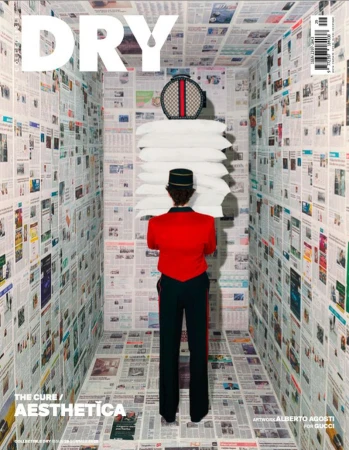 Fashion magazine cover featuring a person in hotel bellhop attire carrying a stack of pillows in a room covered in newspaper.