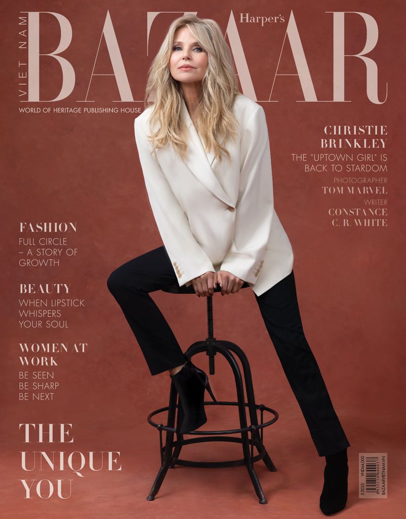 Christie Brinkley poses for Harper's Bazaar Vietnam cover. She's wearing a white blazer, black pants and boots, sitting on a metal stool.