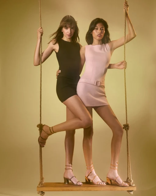 Two adult females pose on a wooden swing suspended by ropes in a studio with diffused lighting, wearing mini dresses and heeled sandals.