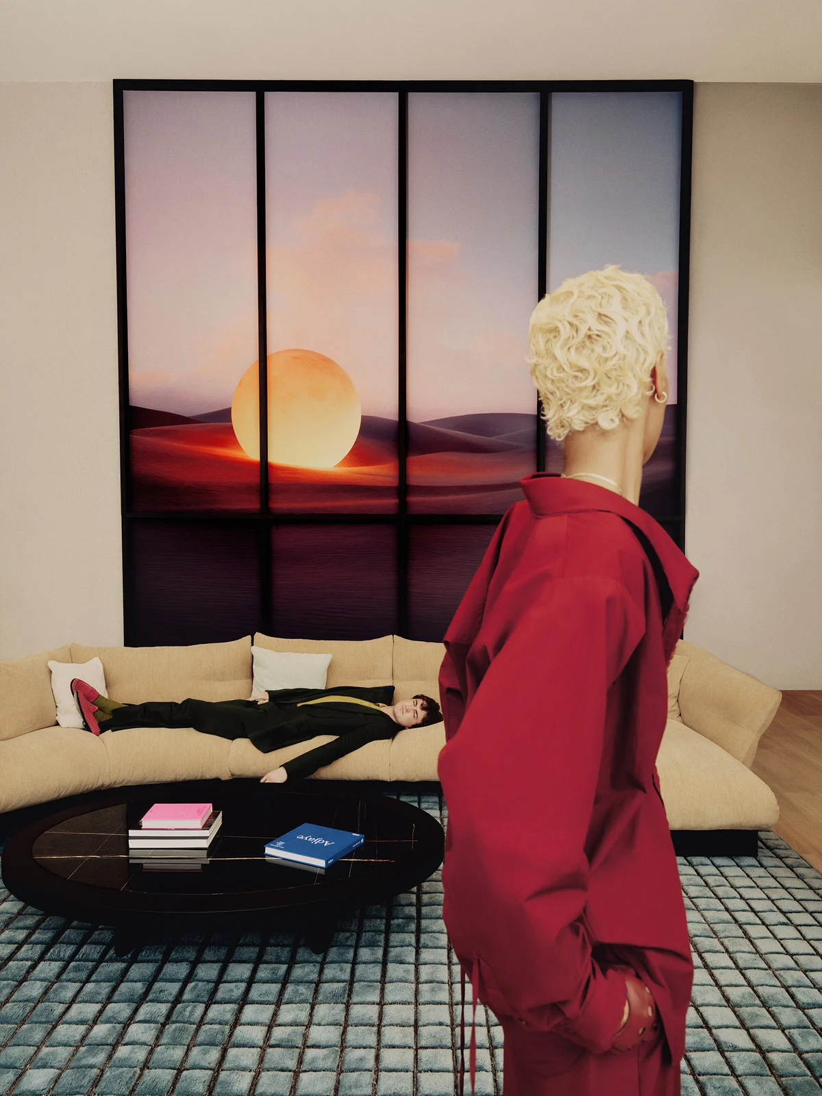 Indoor fashion shot features a person in a red jacket, another lying on a couch, and a blurred person with a large, framed background image.