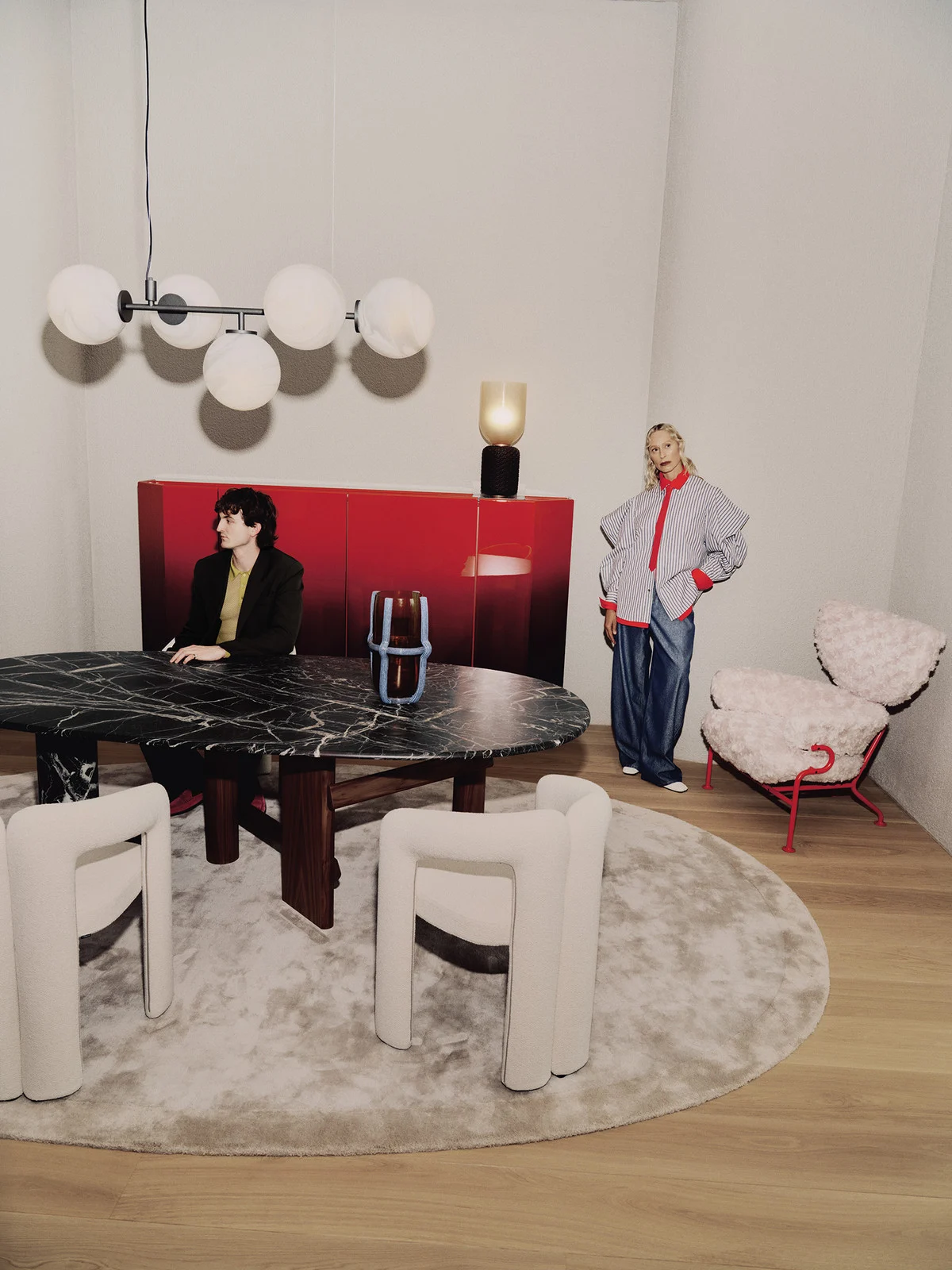 Interior fashion shot of two adults in a room with a marble table, unique chairs, round rug, modern lighting and red sideboard.