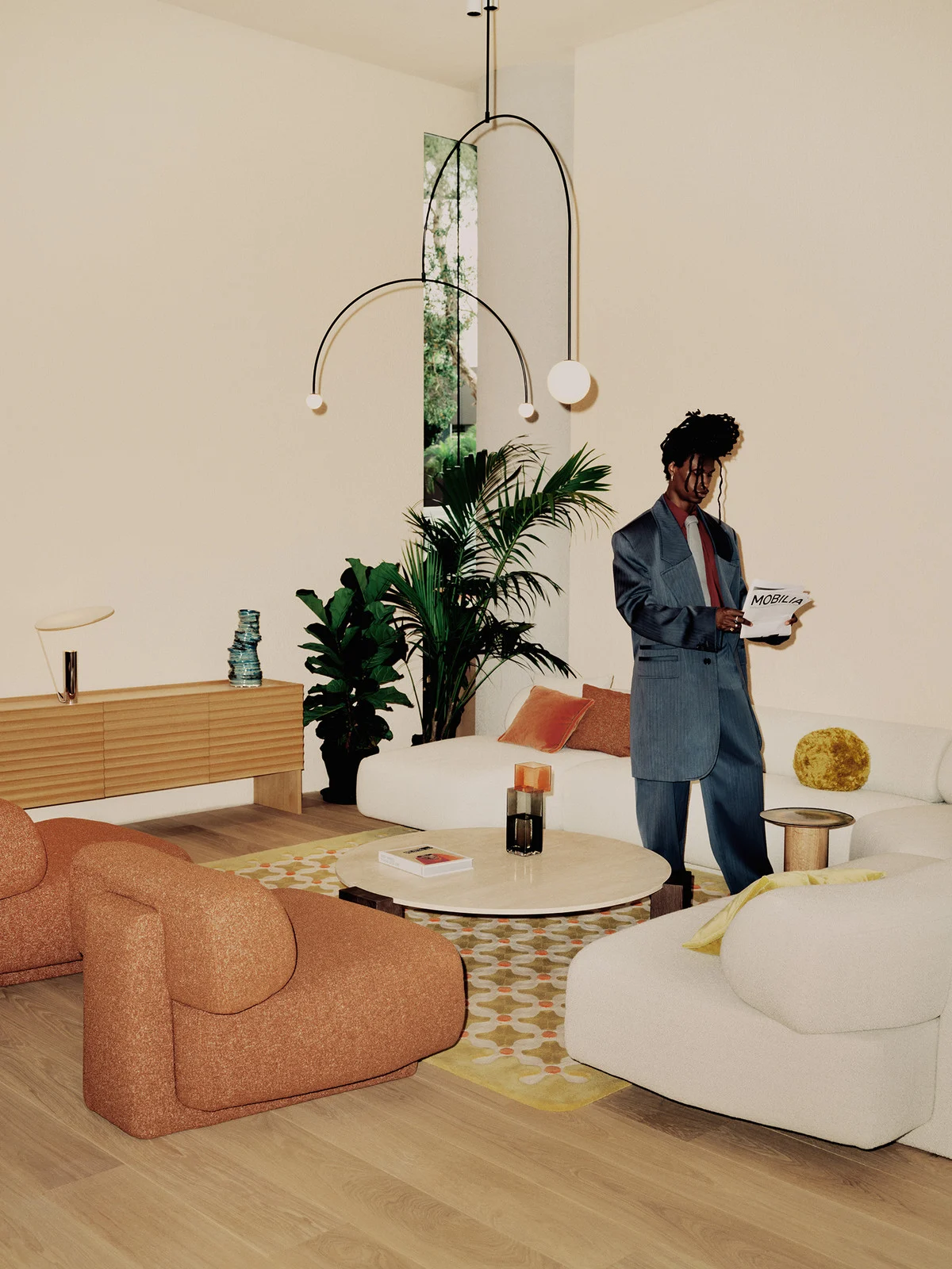 A man in a blue suit stands in a well-lit living room holding a newspaper, flanked by modern furniture and indoor plants creating a stylish scene.