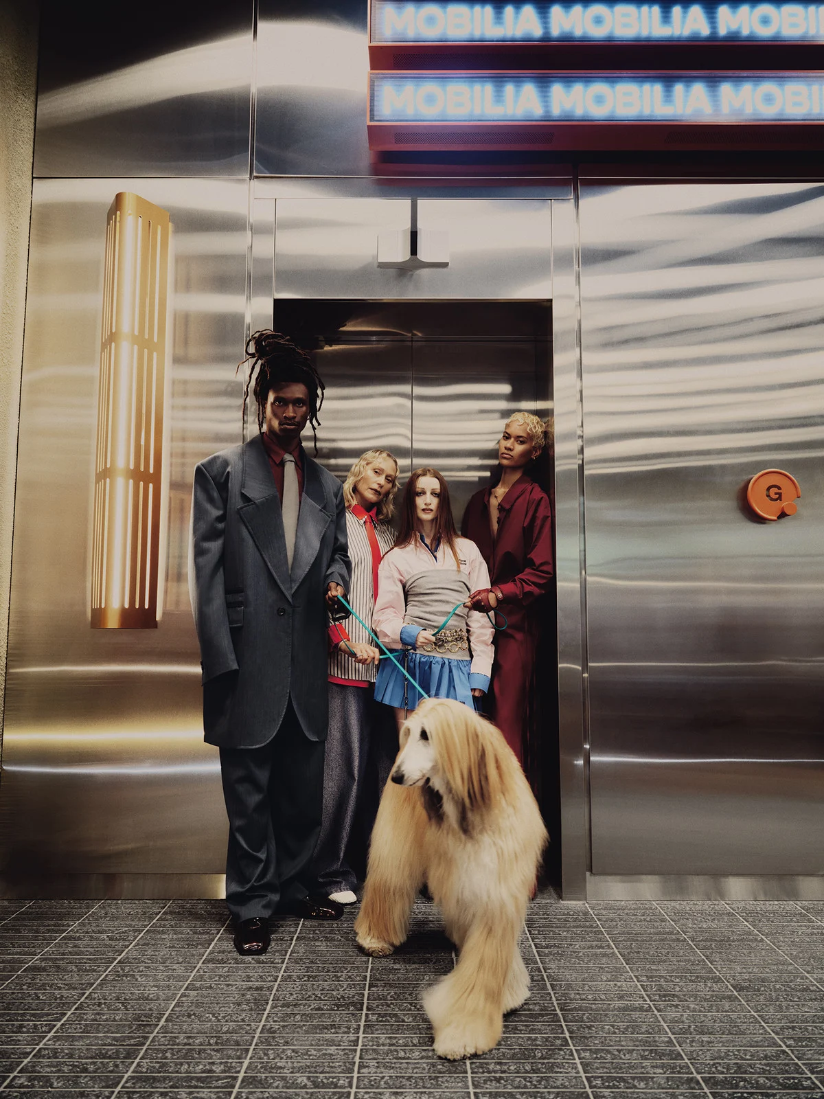Group of stylishly dressed adults and an Afghan Hound dog pose inside a building in front of brushed silver elevator doors and neon signage.