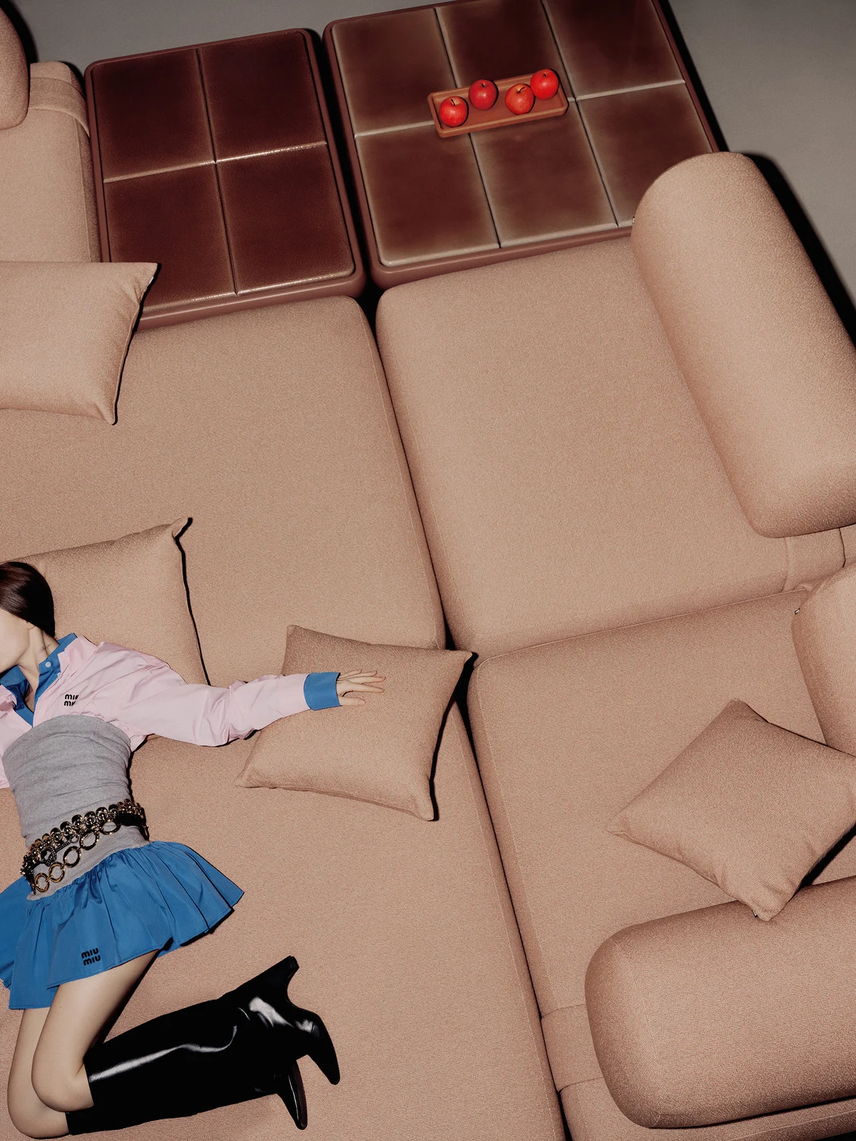 Young woman lounges on a peach-colored sectional sofa in a fashion-style photo. She wears a blue skirt, black boots, and silver accessories.