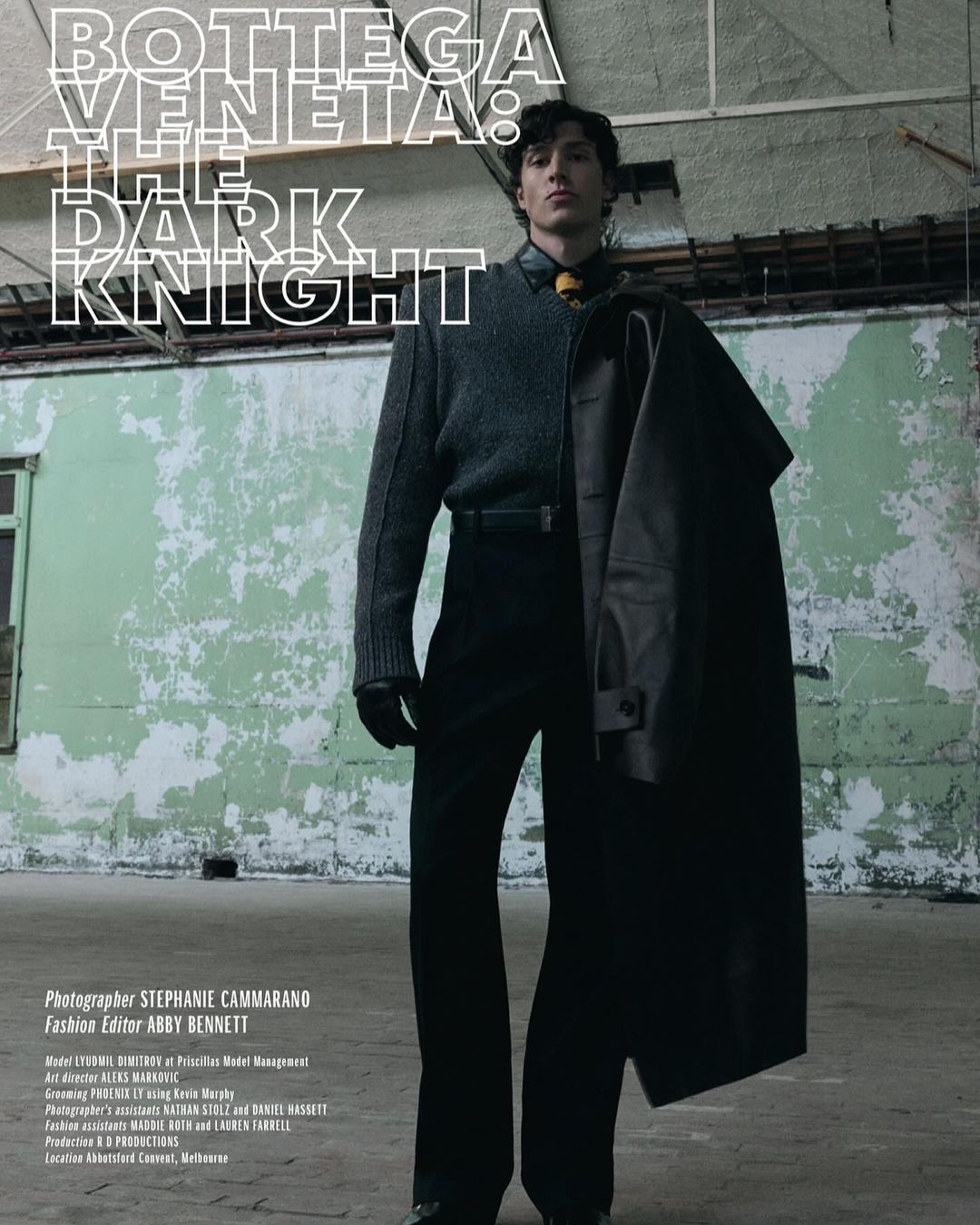 A man in black pants and a grey sweater holds a black coat in an industrial setting, with the words Bottega Veneta in the background.