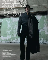 A young male model in dark trousers and sweater standing in an industrial space with distressed green walls holding a leather coat