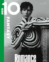 A young man with curly dark hair wearing a patterned striped sweater poses against a plain studio background.