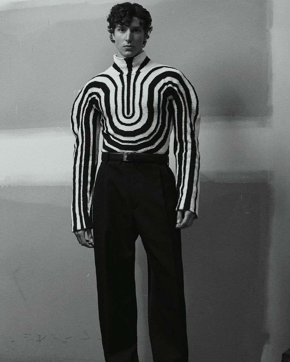 Man wearing a black and white striped patterned turtleneck sweater and dark trousers against a plain studio background.