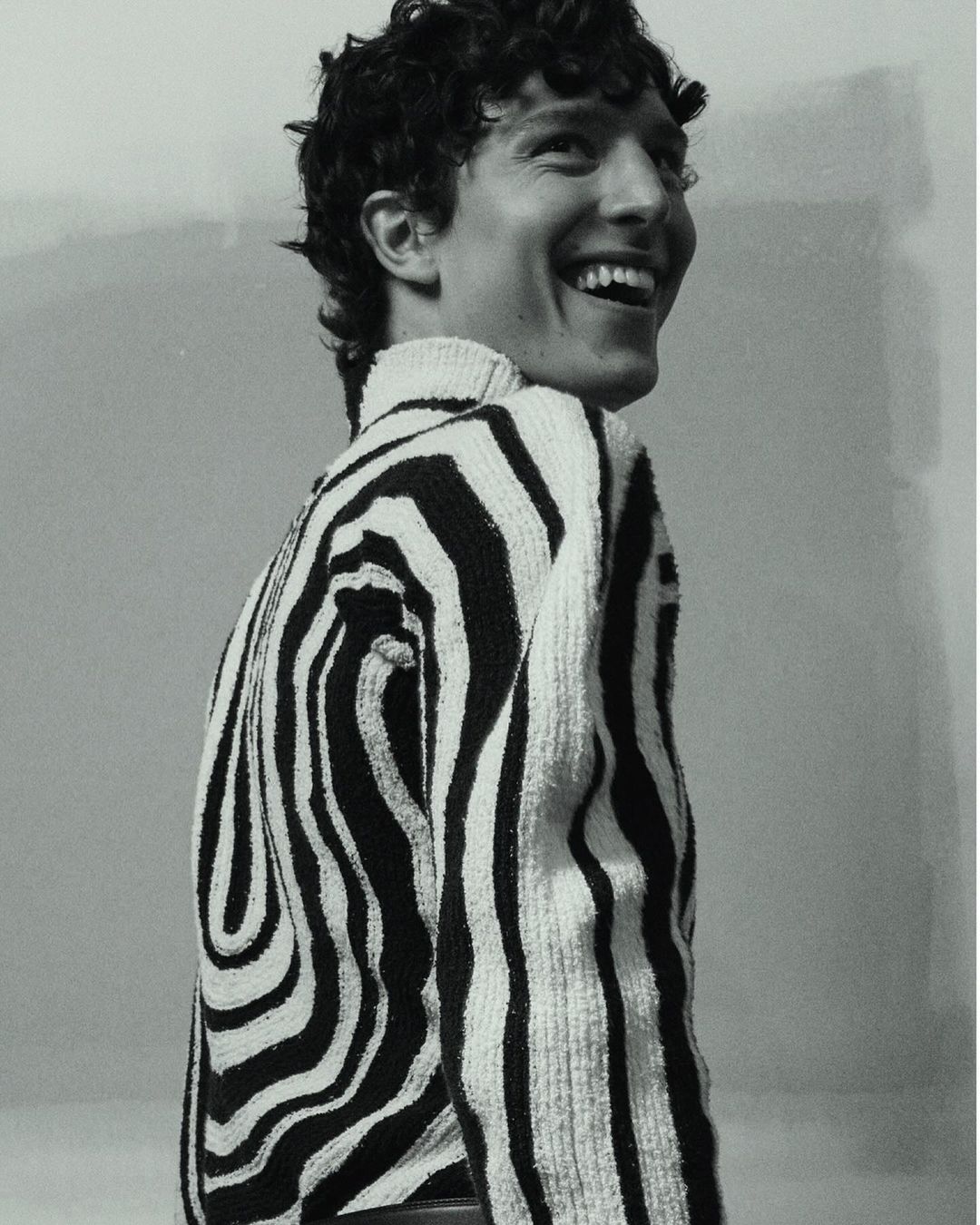 Smiling adult male in zebra-striped sweater stands indoors against neutral wall in close-up studio shot, showcasing fashion and style.