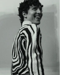 A smiling young man with dark curly hair wearing a black and white striped turtleneck sweater against a plain background.