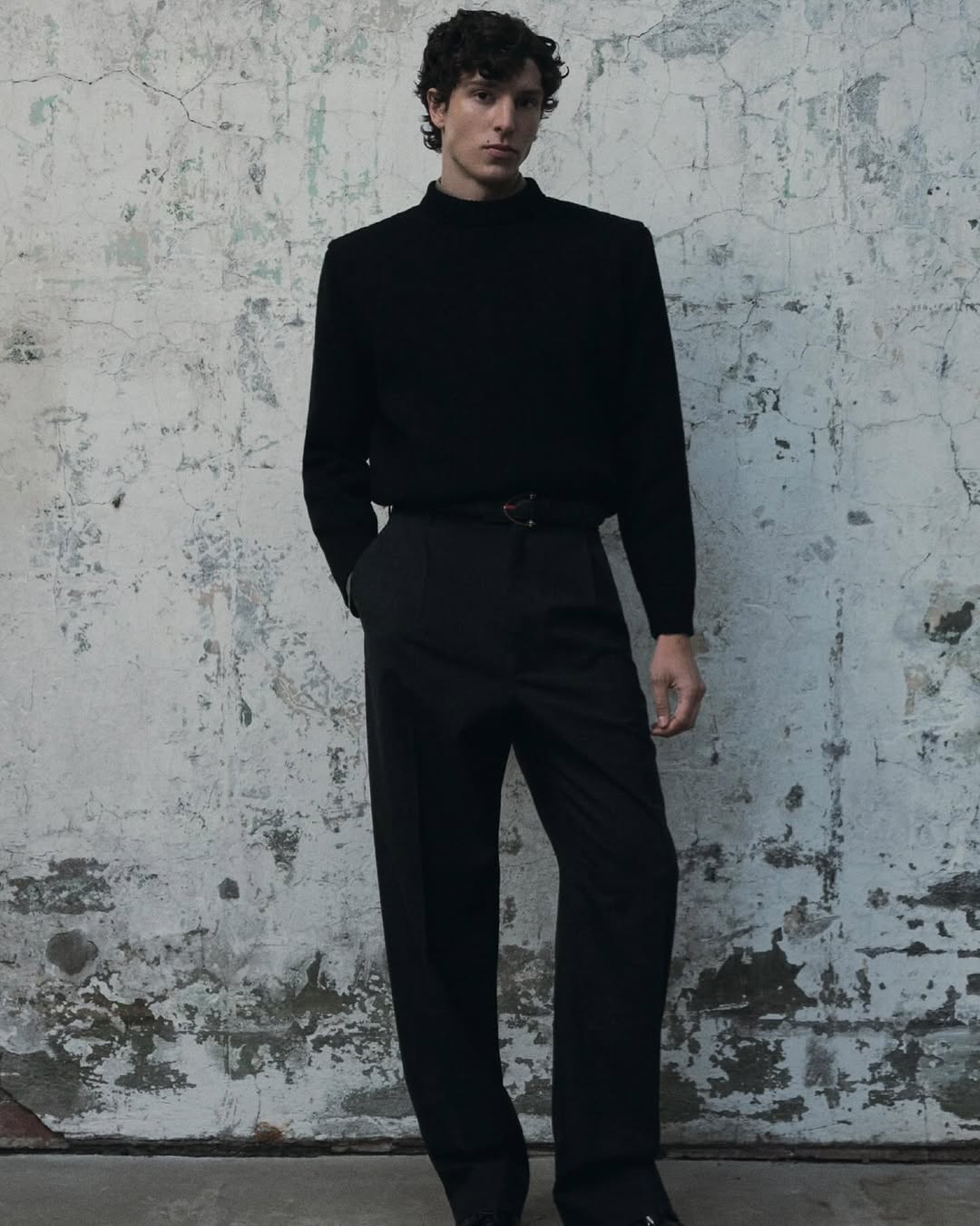 Man wearing a black turtleneck and black trousers standing against a weathered concrete wall for 10 Magazine