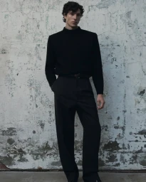 Man wearing a black turtleneck and black trousers standing against a weathered concrete wall for 10 Magazine