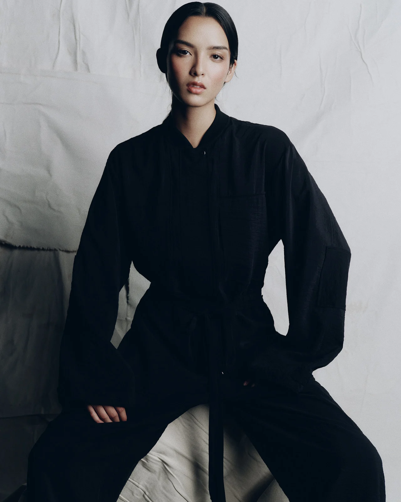 An adult woman poses in a black outfit, sitting against a textured white backdrop in a fashion or portrait photo, styled with clean lines.