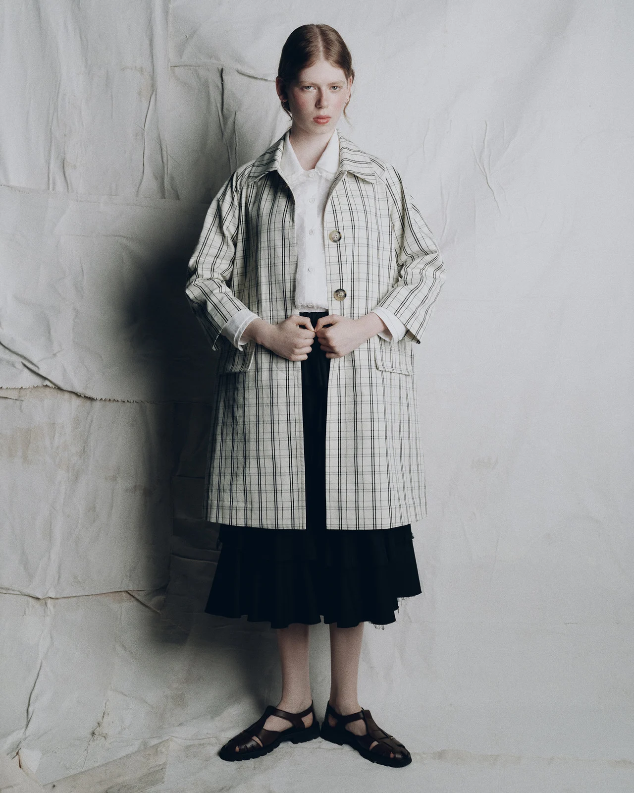 Full-length shot of a young adult woman in a plaid coat and sandals against a textured white background. Fashion editorial portrait.