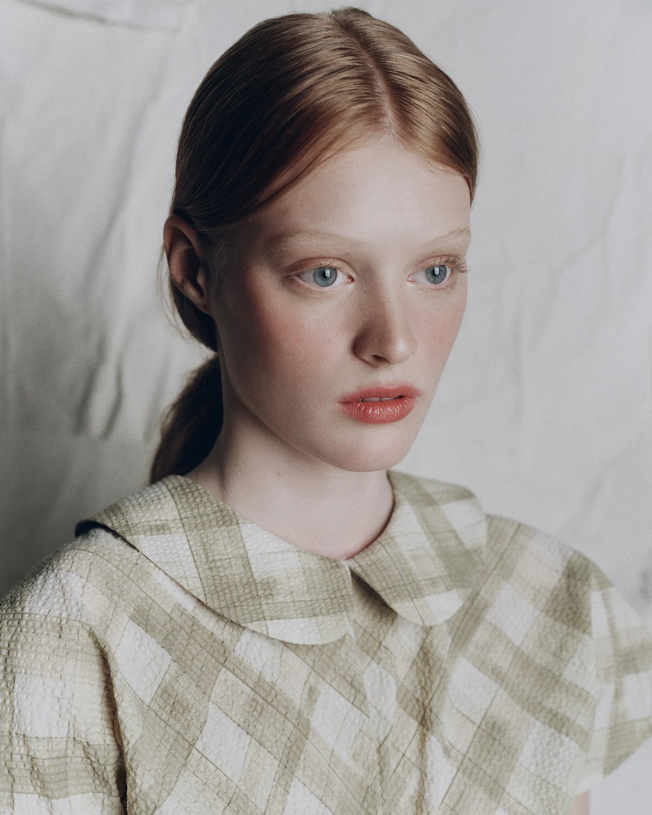 Portrait of a fair-skinned young woman with red hair wearing a light green and white checkered top against a textured white background.