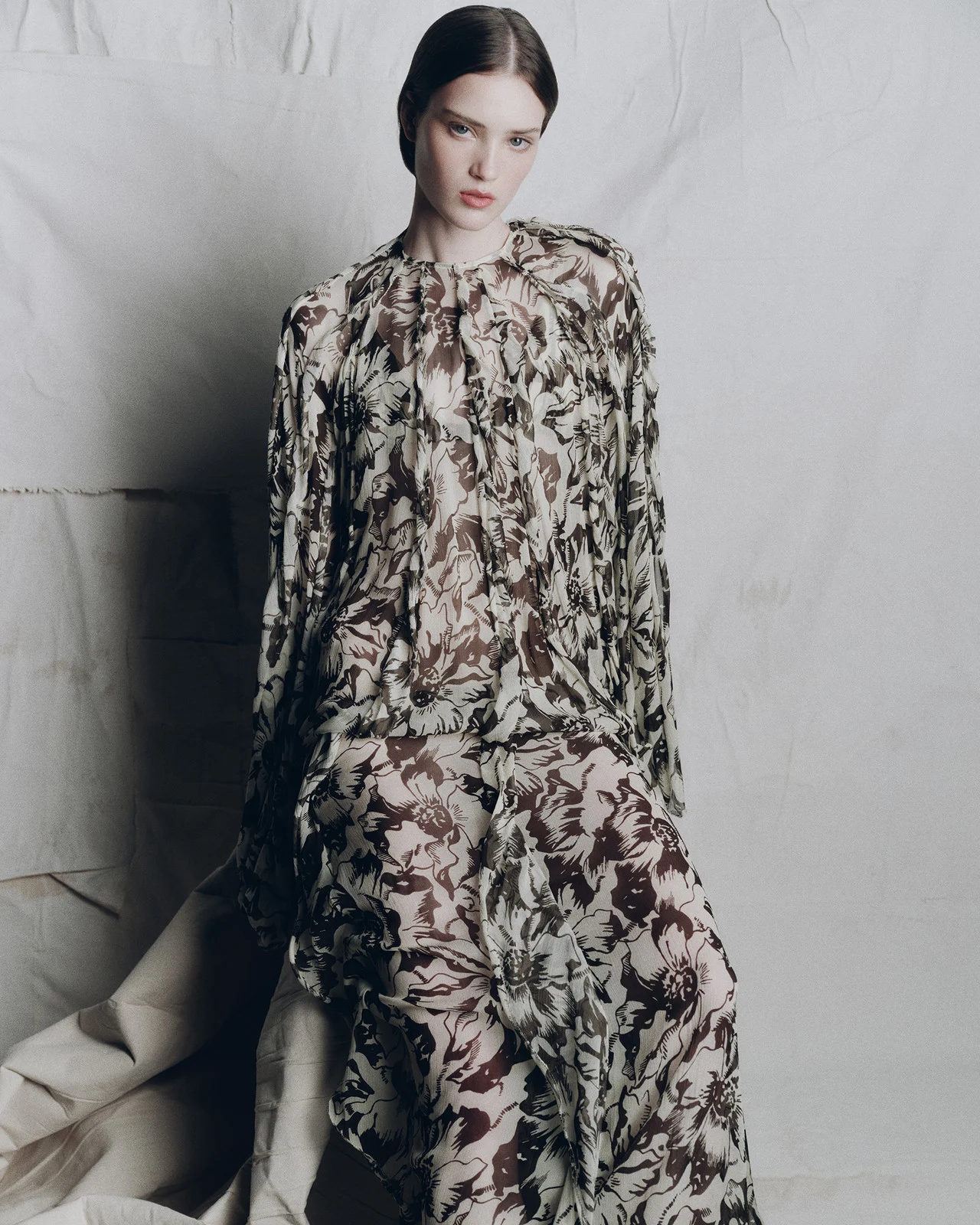A fair-skinned adult woman is posing in a floral print dress, seated against a textured beige background in a fashion editorial shot.