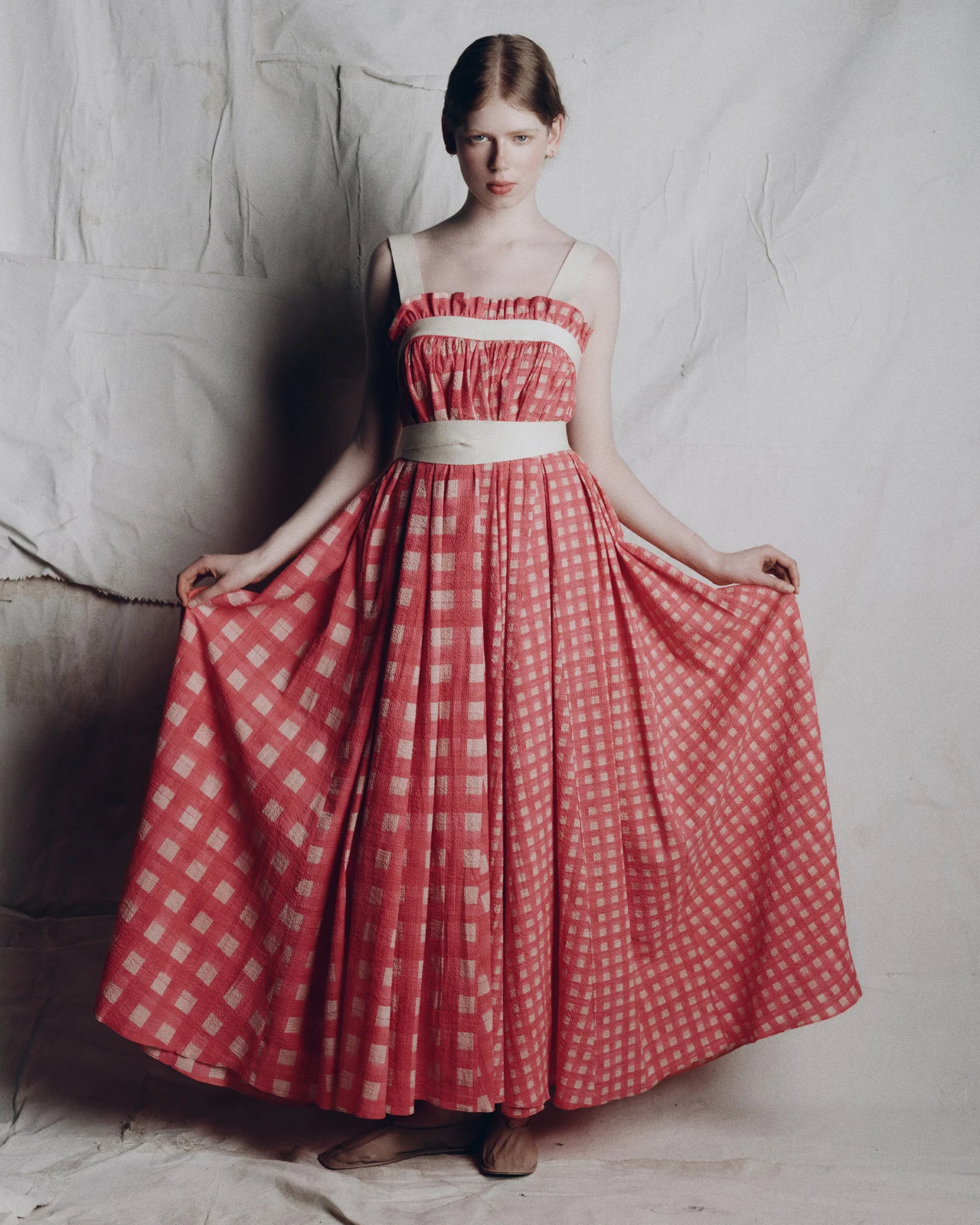 Full shot of a young fair-skinned woman in a long red and white checkered dress, posing in front of a wrinkled white backdrop.