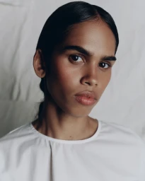 Close-up portrait of a woman with dark hair tied back, wearing a white top against a textured white background.