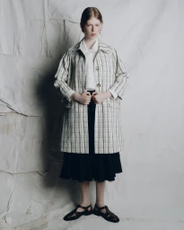 Model wearing a plaid coat over a white shirt and black skirt against textured backdrop