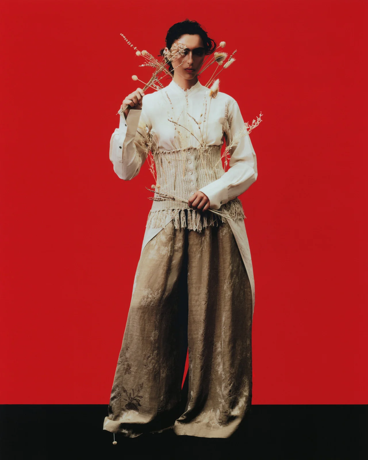 Full-length portrait of a female model in a cream corset, dress shirt, and floral pants, holding dried flowers against a red backdrop.