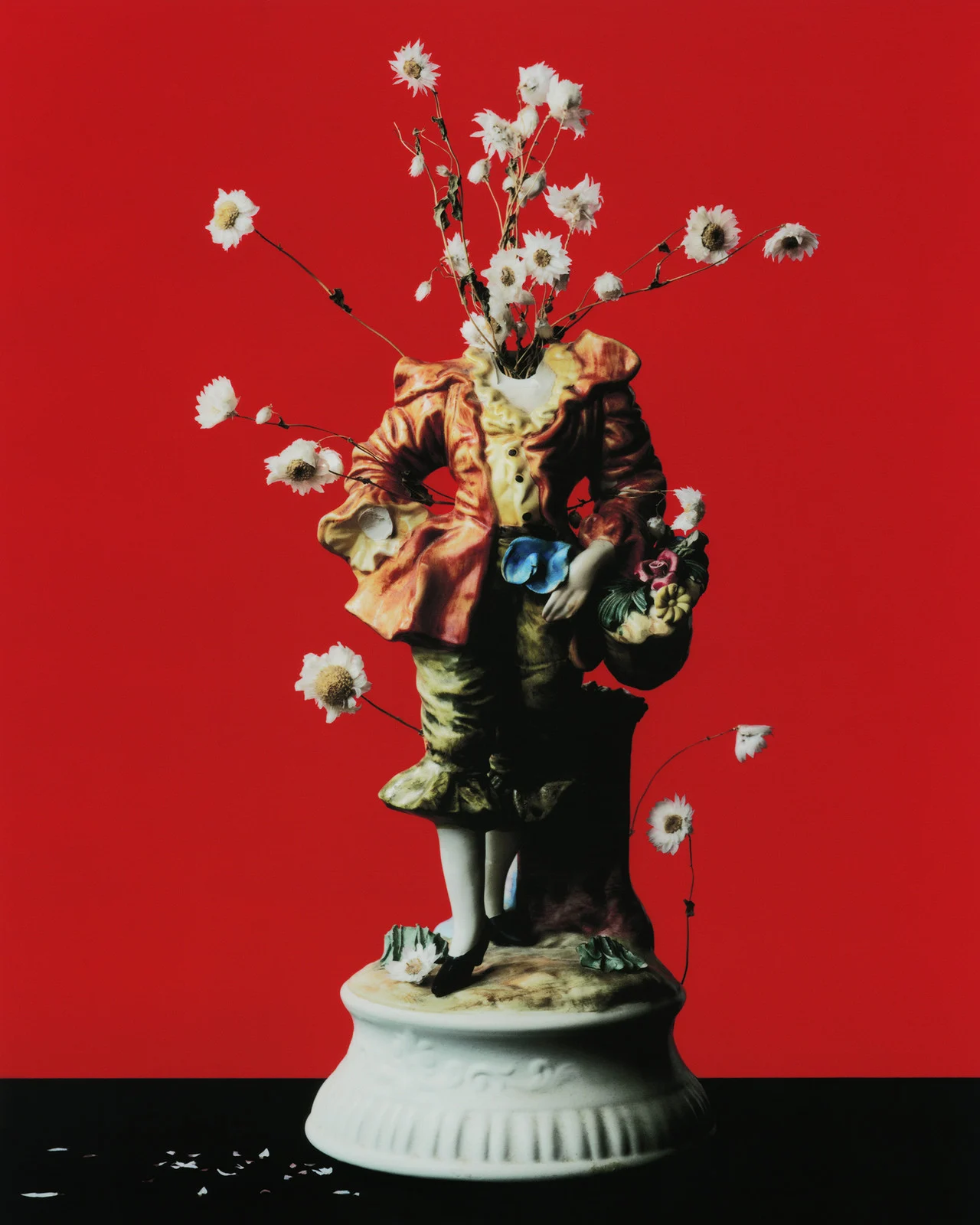 Ornate porcelain figurine on a black surface against a red backdrop, adorned with daisy-like flowers sprouting from the figurine's neck.