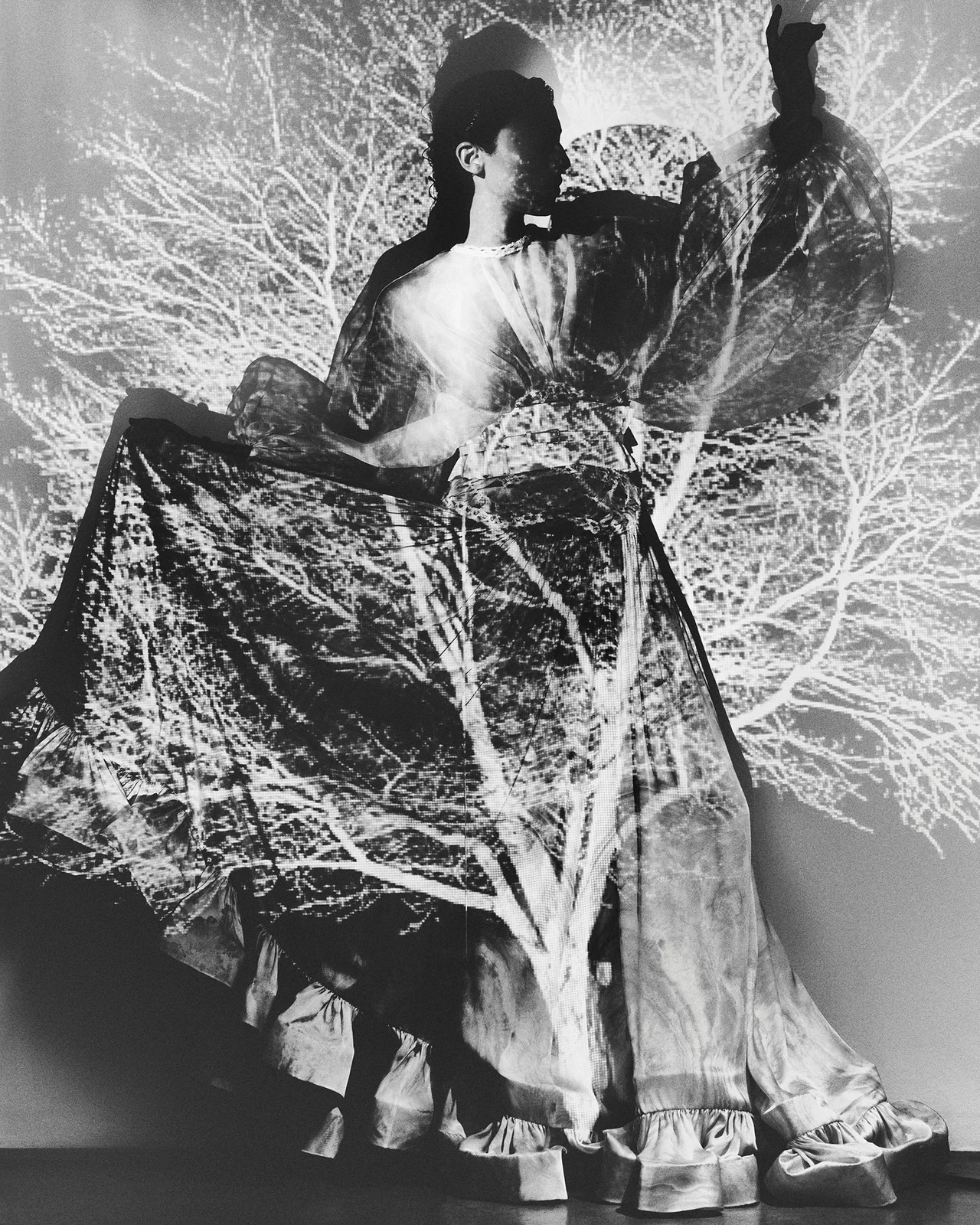 Monochrome double exposure of a woman in flowing dress, superimposed with bare tree branches creating a ghostly, surreal composition.