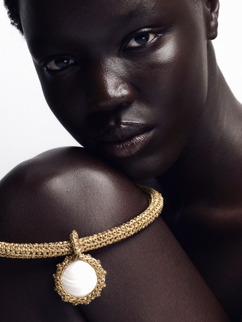 Close-up studio portrait of a Black woman with a neutral expression wearing a gold woven necklace with a shell pendant on a white background.