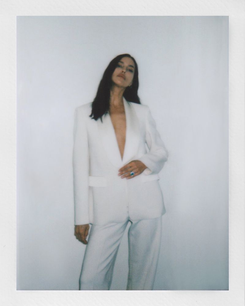 A stylish woman with long dark hair poses in a white pantsuit against a white background in a soft-focus indoor fashion shot.