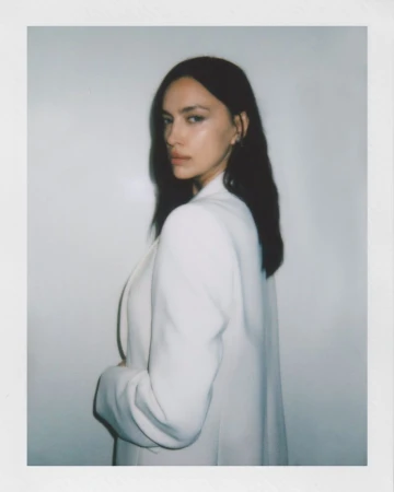 Portrait of a woman with dark hair in a white blazer against a neutral background, looking over her shoulder at the camera.