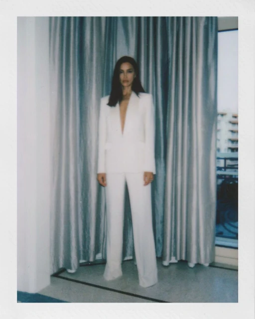 Adult female poses indoors wearing a white suit. The background features neutral curtains and buildings, creating a professional fashion shot.
