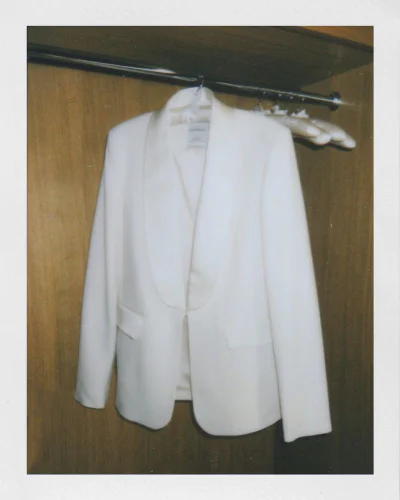 White blazer hanging on a metal rod inside a wooden wardrobe with a Polaroid-style white border.