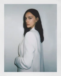 Model Irina Shayk wearing a white blazer against a white seamless background in a polaroid portrait.