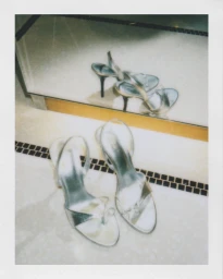Polaroid photograph of silver high-heeled slingback sandals reflecting in a mirror on a marble tile floor