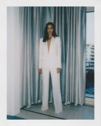 Model Irina Shayk wearing a white pantsuit standing against grey curtains inside a hotel room
