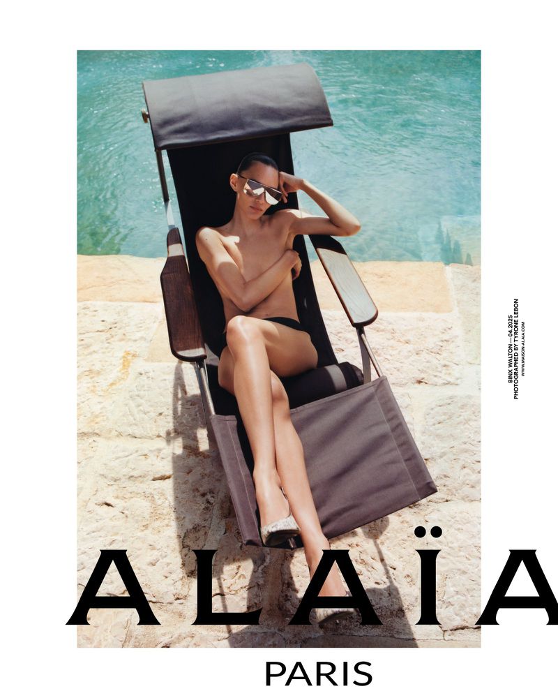 Fashion shot of a slender, light-skinned woman in sunglasses and black swimwear, reclining on a beach chair near a pool. Alaïa branding.