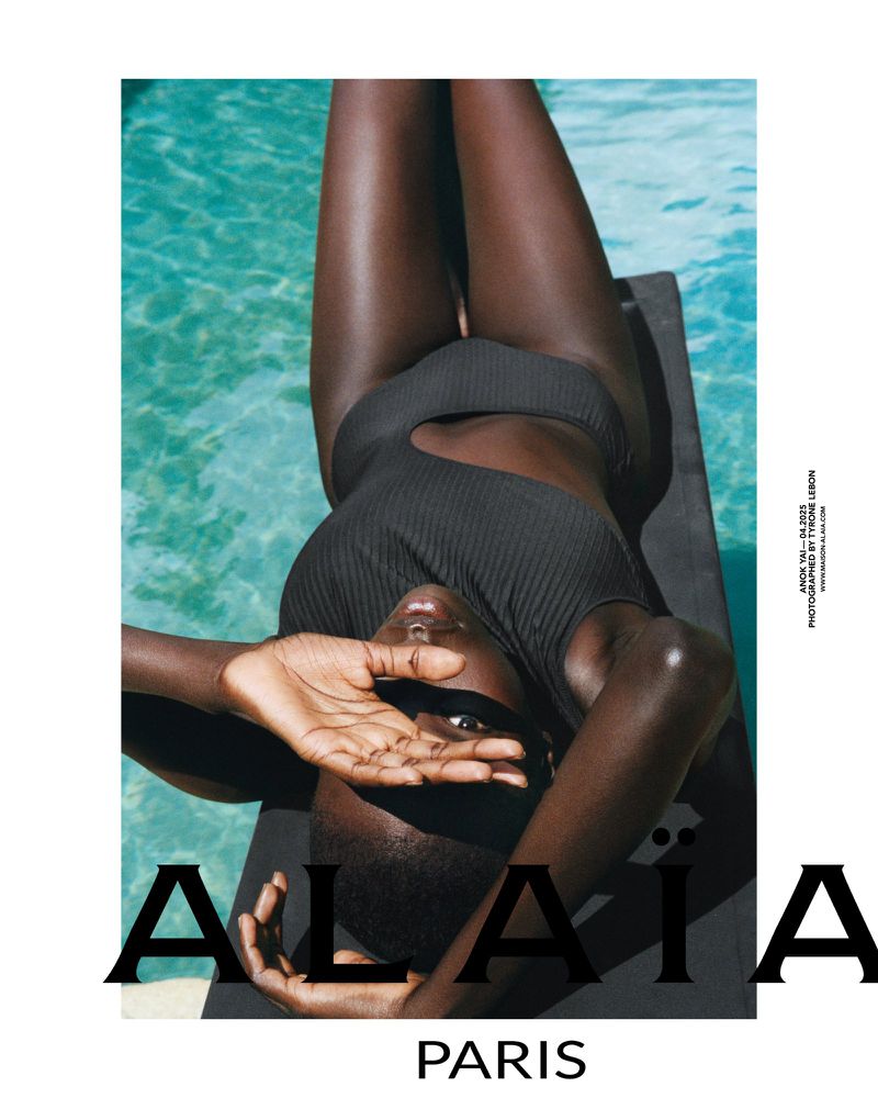 A woman in a black swimsuit lies on a dark surface by a pool with turquoise water. Her hand shields her eyes from the light.