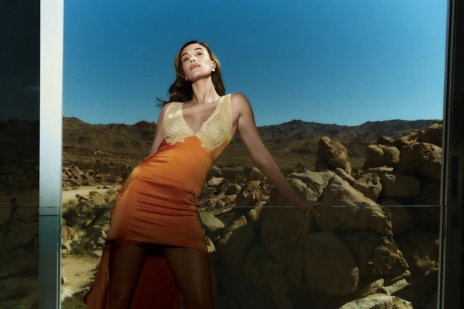 Woman poses in a dress against a desert backdrop with mountains. She leans on rocks, under a blue sky, reflecting light.