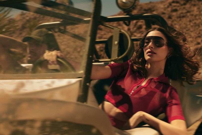 A woman with sunglasses driving a beige off-road vehicle in a desert landscape, with a man visible as a passenger.