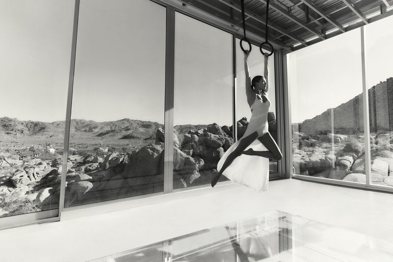 Female gymnast performs on rings in a modern architectural interior with desert landscape backdrop, captured in a black and white photograph.