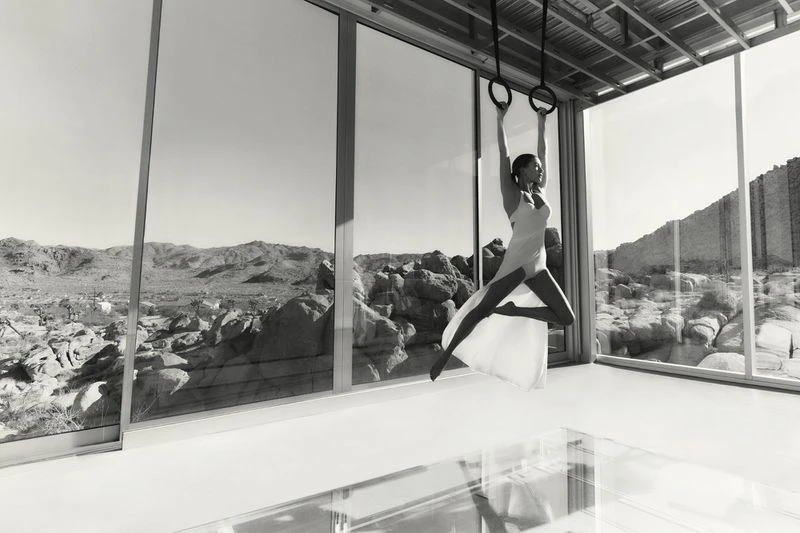 Female gymnast performs on rings in a modern architectural interior with desert landscape backdrop, captured in a black and white photograph.