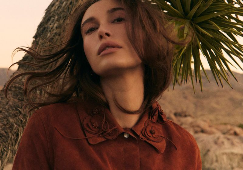 Close-up of a young adult female fashion model with windblown brown hair, wearing a rust-colored shirt with a floral collar in an outdoor setting.