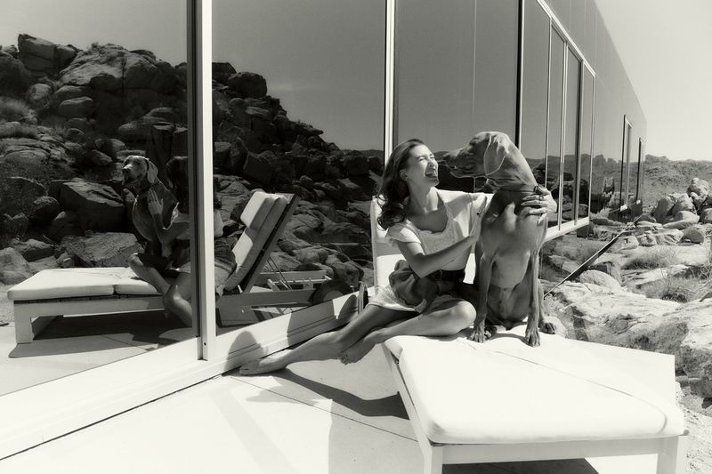 A woman sits on a white lounge chair with a dog on a patio. Rock formations and a modern building are in the background.