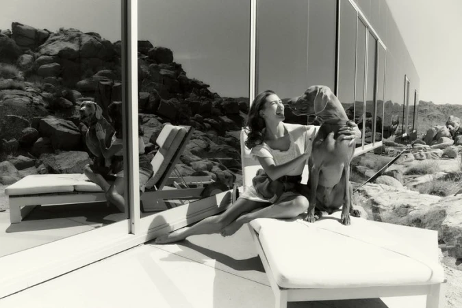 A woman sits on a white lounge chair with a dog on a patio. Rock formations and a modern building are in the background.