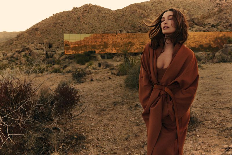 Adult female posing in a brown dress and robe in desert landscape with hills and copper-colored mirrored architectural building.