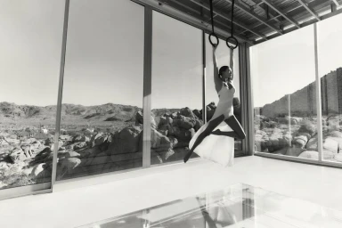 Model in a white dress suspended from gymnastic rings inside a modern glass house overlooking a rocky desert landscape.