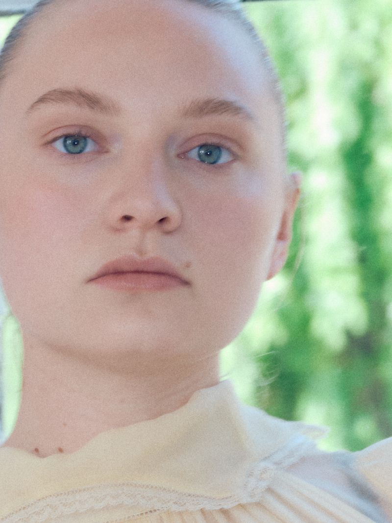 Close-up portrait of a young woman with blue eyes and pale skin, wearing a light yellow top with lace details against a blurred green background.