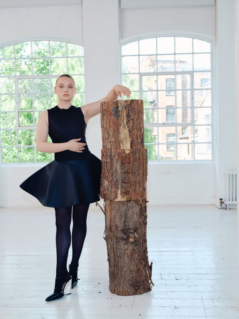 A young woman in a black dress and dark tights leans on stacked logs in a white photo studio with large windows. Fashion editorial.